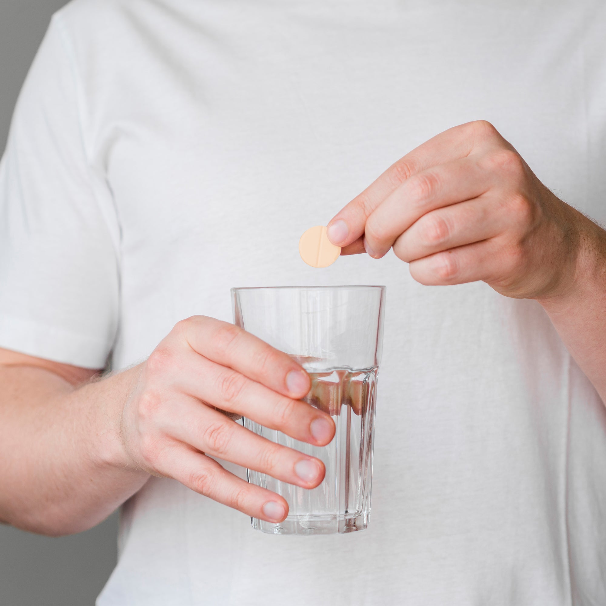 Person holding a glass of water and a pill against a neutral background